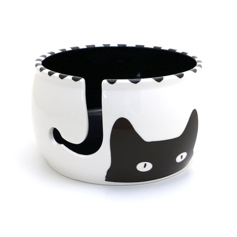 May include: White ceramic yarn bowl with a black cat design. The cat's face is visible with one eye and part of the other eye. The cat's tail is the yarn holder.
