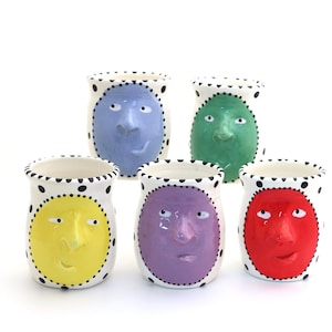 May include: Five ceramic face vases in various colors: yellow, purple, red, green, and blue. Each vase has a white base with black polka dots around the rim and a face design in the center.