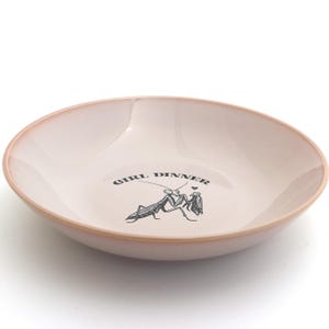 Peut inclure: Un bol en c&eacute;ramique rose clair avec un bord l&eacute;g&egrave;rement relev&eacute;. Le bol porte l'inscription "GIRL DINNER" et une illustration noire de deux mantes religieuses. Id&eacute;al pour servir des aliments.