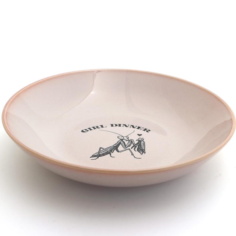 Peut inclure: Un bol en c&eacute;ramique rose clair avec un bord l&eacute;g&egrave;rement relev&eacute;. Le bol porte l'inscription "GIRL DINNER" et une illustration noire de deux mantes religieuses. Id&eacute;al pour servir des aliments.