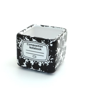 May include: A square ceramic planter featuring a black and white speckled pattern reminiscent of a classic composition notebook. The front displays a white label with the printed text Composition Notebook and two horizontal lines for writing.