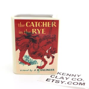 May include: A ceramic book-shaped vase with the title "The Catcher in the Rye" on the front. The cover art features a red illustration of a person on a horse. The vase is off-white with a rectangular shape.