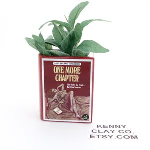 May include: A red ceramic planter shaped like a book with the title "One More Chapter" and the text "Just a girl who loves books" and "She woke up tired... But also smarter". The planter has a green plant growing out of the top.