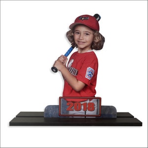 May include: A young girl in a red baseball uniform holds a blue and white baseball bat. She is wearing a red hat with a white owl logo and the year 2018 is displayed on a plaque below her.