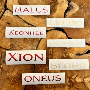 Puede incluir: Siete pegatinas rectangulares blancas con los nombres "Malus", "Leedo", "Keonhee", "Hwanwoong", "Xion", "Seohyo" y "Oneus" impresos en letras rojas y doradas.