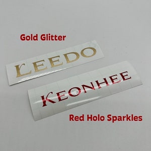 Puede incluir: Dos pegatinas blancas con nombres en ellas. La pegatina superior tiene el nombre "LEEDO" en letras doradas brillantes. La pegatina inferior tiene el nombre "KEONHEE" en letras hologr&aacute;ficas rojas. El texto "Gold Glitter" est&aacute; por encima de la pegatina superior y el texto "Red Holo Sparkles" est&aacute; por debajo de la pegatina inferior.