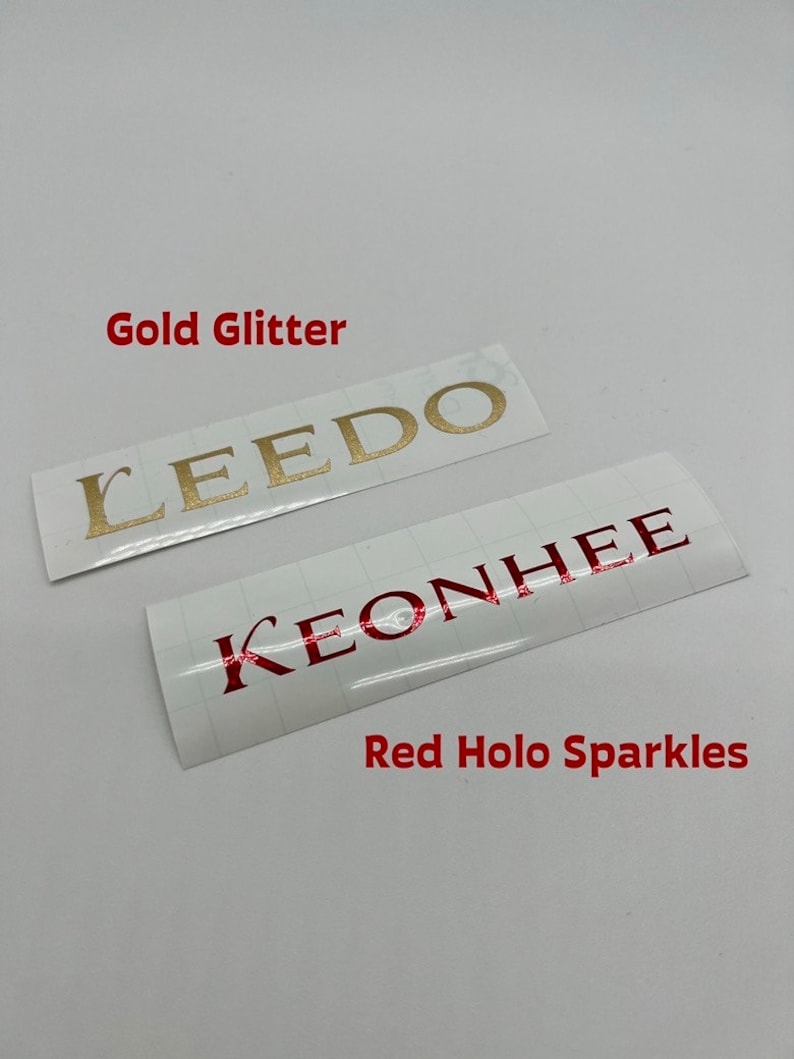 Puede incluir: Dos pegatinas blancas con nombres en ellas. La pegatina superior tiene el nombre "LEEDO" en letras doradas brillantes. La pegatina inferior tiene el nombre "KEONHEE" en letras hologr&aacute;ficas rojas. El texto "Gold Glitter" est&aacute; por encima de la pegatina superior y el texto "Red Holo Sparkles" est&aacute; por debajo de la pegatina inferior.