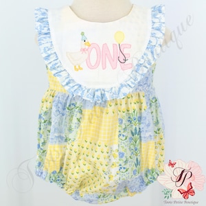 May include: A yellow, blue, and white patchwork baby onesie with a white bib featuring a pink embroidered "ONE" and a cartoon duck wearing a party hat.