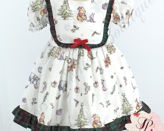 May include: White dress with a Winnie the Pooh Christmas print. The dress features short, puffed sleeves, a red velvet bow, and green and red plaid trim. The dress has a ruffled hem and is designed for a child.