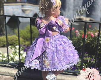 May include: A lavender princess-style dress with a matching headband. The dress features a large bow in the back and is adorned with floral and sequin embellishments. The dress is worn with white shoes.