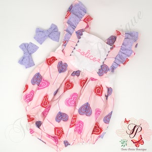 May include: A pink baby romper with a heart and lollipop pattern, featuring the word "sweet" and "oxox". The romper has ruffled sleeves and trim, with the name "alice" embroidered on the front. Two matching lavender polka dot bows are included.