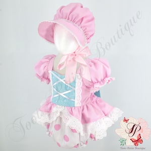 May include: A pink baby outfit with a matching bonnet. The outfit features a light blue bodice with white criss-cross ribbon detail, pink puff sleeves, and a ruffled skirt over white polka dot bloomers. The bonnet has a wide brim and lace trim.