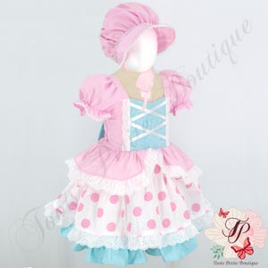 May include: A pink and light blue dress with a matching bonnet. The dress features a light blue bodice with white crisscross ribbon detailing, pink puff sleeves, and a white skirt with large pink polka dots, finished with white eyelet lace trim.