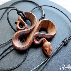 May include: A wooden snake pendant, intricately carved and stained a rich brown. The pendant hangs from a black cord. The snake's head and body are detailed. The pendant is displayed on a dark surface. The artist's name is visible in the image.