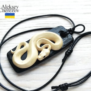 Könnte beinhalten: Eine schwarze Halskette mit einem rechteckigen schwarzen Anhänger, in den eine weiße Schlange geschnitzt ist. Die Halskette besteht aus schwarzem Kordel.