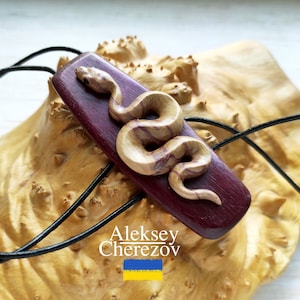 May include: A wooden snake pendant with a purple and brown wood grain pattern. The pendant is on a black leather cord.