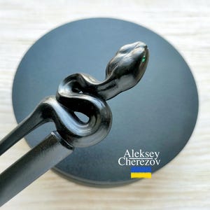 Könnte beinhalten: Eine schwarze Haargabel mit Schlangenmuster. Der Schlangenkopf befindet sich oben, mit einem kleinen grünen Akzent. Die Gabel befindet sich vor einem dunkelgrauen kreisförmigen Hintergrund. Der Name des Künstlers und eine ukrainische Flagge sind sichtbar.