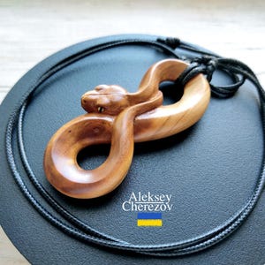 May include: A wooden pendant necklace featuring a carved snake in a figure-eight design. The snake is light brown with detailed carving, and the necklace has a black cord. The artist's name and a Ukrainian flag are visible.