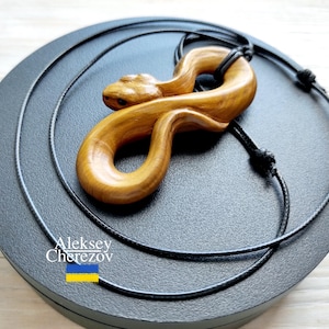 May include: A wooden snake pendant necklace. The pendant is carved from light brown wood and features a detailed snake design. The necklace has a black cord and is displayed on a black surface. The artist's name, Aleksey Cherezov, is visible.