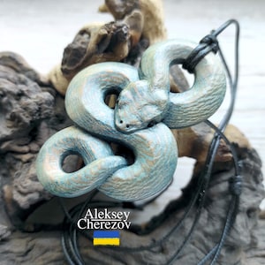 May include: A coiled snake pendant with a distressed teal and bronze finish. The pendant is suspended from a black cord. The artist's name, Aleksey Cherezov, is visible in the image, along with the Ukrainian flag.