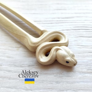 Könnte beinhalten: Eine hellfarbene Haar-Gabel aus Holz, die in Form einer Schlange geschnitzt ist. Der Schlangenkopf ist mit kleinen blauen Augen detailliert. Die Gabel hat zwei Zinken und der Name des Künstlers, Aleksey Cherezov, ist sichtbar.