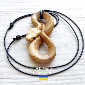 May include: A wooden snake pendant necklace. The pendant is carved from light-colored wood and features a snake design with blue eyes. The snake is coiled in an infinity symbol shape. The necklace has a black cord.