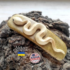 May include: A rectangular hair clip featuring a carved, cream-colored snake with a dark eye, mounted on a smooth, olive-green base. The text Aleksey Cherezov is printed in white, accompanied by a blue and yellow flag and a circular badge.