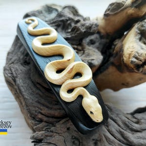May include: A wooden hair clip with a carved snake design. The snake is light brown and the clip is black.