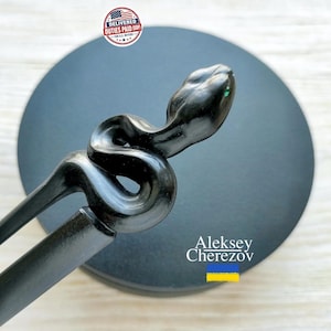 May include: A black hair stick featuring a carved snake head and a coiled body design. The snake is depicted in a smooth, dark finish with a subtle green accent on the head. The accessory has a long, tapered stem extending from the coiled snake.
