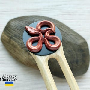 May include: A wooden hair fork with a decorative, reddish-brown snake design on a black circular base. The snake is intricately detailed. The fork is light-colored wood. The name "Aleksey Cherezov" is printed on the bottom left.