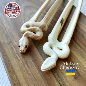 May include: Two wooden hair forks, each featuring a carved snake design. One fork is made of light-grained wood with dark eyes, while the other is a smooth, pale, solid-colored wood. Both items are long, two-pronged accessories for hair styling.
