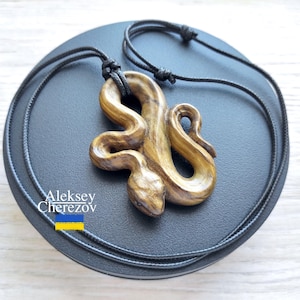 May include: A wooden snake pendant necklace. The pendant, carved in shades of brown and gold, depicts a detailed snake. It hangs from a black cord, displayed on a dark grey surface. The text "Aleksey Cherezov" is visible.