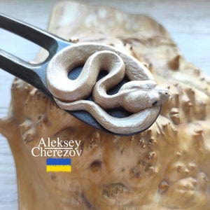 Könnte beinhalten: Eine Holzhaargabel mit einem geschnitzten Schlangenmuster. Die Schlange ist hellbeige und um die Gabel gewickelt. Die Gabel ist schwarz und hat zwei Zinken.