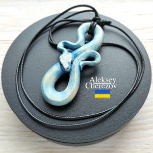 Könnte beinhalten: Eine Halskette mit einem blauen und weißen Schlangenanhänger auf einer schwarzen Schnur. Der Anhänger ist aus Holz geschnitzt und hat ein detailliertes Design. Die Halskette wird auf einer schwarzen runden Box mit dem Namen Aleksey Cherezov und der ukrainischen Flagge präsentiert.
