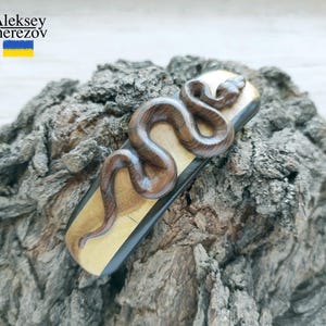 Może przedstawiać: Ręcznie wykonana drewniana figurka węża, misternie wyrzeźbiona i barwiona w odcieniach brązu, spoczywa na prostokątnej podstawie z jasnym usłojeniem drewna. Podstawa jest umieszczona na tle teksturowanej, naturalnej kory. Widoczne jest imię artysty, Aleksey Cherezov.