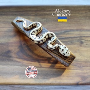 May include: A hair clip featuring a dark brown wood base with a carved, cream-colored snake figure with black markings attached to the top. The text Aleksey Cherezov is printed in white above the snake, accompanied by a small blue and yellow flag.