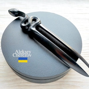 Könnte beinhalten: Eine schwarze Haargabel aus Holz mit Schlangenmuster. Das Haaraccessoire hat einen gewundenen Schlangenkopf oben und zwei lange, spitze Zinken. Die Gabel wird auf einer dunkelgrauen, kreisförmigen Oberfläche präsentiert. Der Text "Aleksey Cherezov" ist sichtbar.