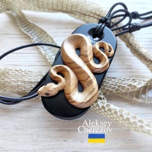 Può includere: Ciondolo a forma di serpente in legno con base nera e cordino nero. Il serpente è scolpito in legno marrone chiaro con venature marroni più scure. Il ciondolo è su una superficie di legno chiaro con pelle di serpente.