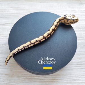 Może przedstawiać: Drewniana figurka węża z kremowo-brązowym wzorem, spoczywająca na ciemnoszarej okrągłej podstawie. Podstawa zawiera napis "Aleksey Cherezov" oraz niebiesko-żółtą flagę.