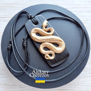 Może przedstawiać: Drewniany naszyjnik z wisiorkiem przedstawiającym złotego węża zwiniętego na czarnej prostokątnej podstawie. Wąż ma szczegółowy wzór i małe zielone oko. Naszyjnik ma czarny sznurek. Widoczne jest imię artysty.