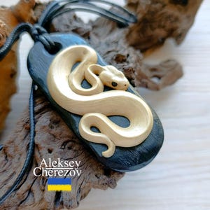 Può includere: Un ciondolo nero con un design a forma di serpente dorato. Il ciondolo è su un cordoncino nero e pende da un pezzo di legno galleggiante. Il testo "Aleksey Cherezov" è visibile sul legno galleggiante.