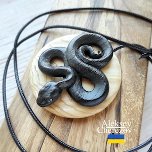 May include: Black snake pendant necklace on a black cord. The snake is carved in wood and is coiled on a round wooden base. The pendant is on a wooden surface with the text "Aleksey Cherezov" and a Ukrainian flag.