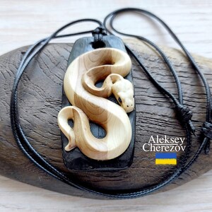 Peut inclure: Pendentif en bois représentant un serpent sculpté. Le serpent est en bois clair, posé sur une base rectangulaire foncée. Un cordon noir est attaché pour le porter. Un bijou unique.