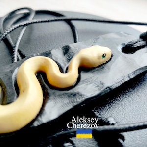 May include: A handcrafted pendant necklace featuring a cream-colored snake figurine with green eyes, resting on a black, textured tag. The pendant is suspended from a black cord. The artist's name, Aleksey Cherezov, is visible in the image.