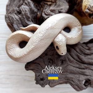 Op de afbeelding: Een wit houten slangenbeeldje dat in een opgerolde positie is gesneden. Het beeldje rust op een stuk donkerbruin drijfhout. De naam van de kunstenaar, Aleksey Cherezov, staat onder het beeldje, samen met de Oekraïense vlag.