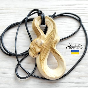 May include: A hand-carved wooden pendant shaped like a coiled snake with a light tan and brown wood grain pattern, suspended on a thin black cord. The pendant features small dark eyes and a smooth finish. Text reads Aleksey Cherezov with a flag.