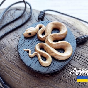 May include: A wooden snake pendant necklace with a black cord. The snake is carved in detail and has a natural wood color. The pendant is round and has a black background.