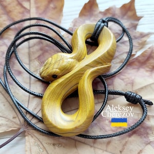 Op de afbeelding: Een gesneden gele houten slangen hanger in de vorm van een doorlopende lus, hangend aan een zwart koord. De houtnerf is zichtbaar over het gehele gladde oppervlak van de slang. De tekst Aleksey Cherezov staat gedrukt bij een kleine blauwe en gele vlag.