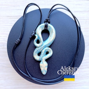 Może przedstawiać: Naszyjnik z niebiesko-białą zawieszką w kształcie węża z czarnym sznurkiem. Zawieszka w kształcie węża jest misternie zaprojektowana ze zwiniętym ciałem i szczegółową głową. Naszyjnik jest prezentowany na ciemnoniebieskiej powierzchni.