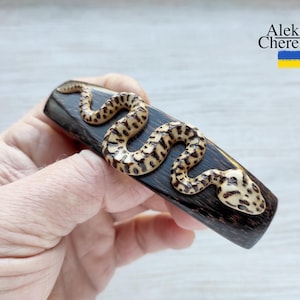 May include: A wooden hair clip with a carved snake design. The snake is light brown and black with a detailed pattern. The clip is dark brown and has a smooth finish.